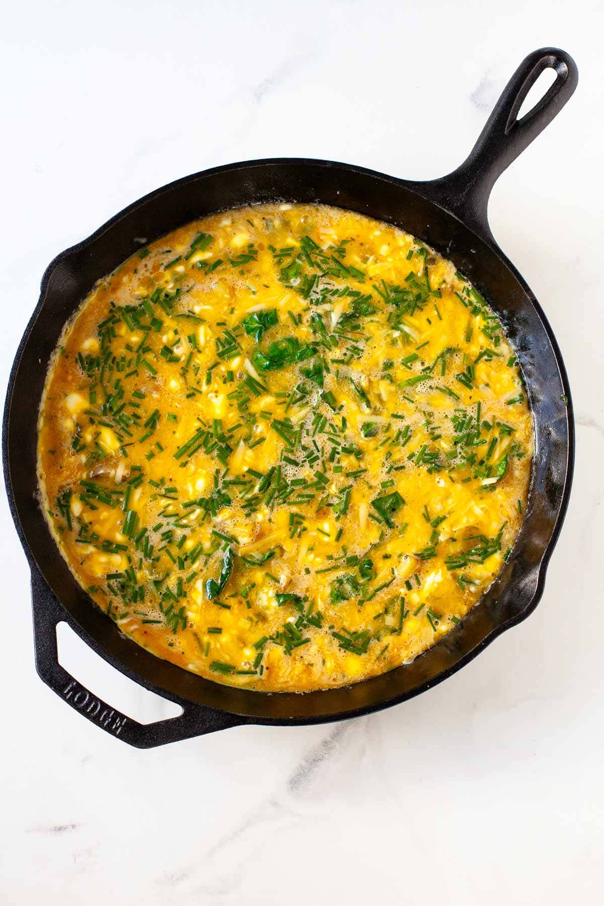 partially cooked swiss chard potato chive frittata in black cast iron pan with edges set on marble surface