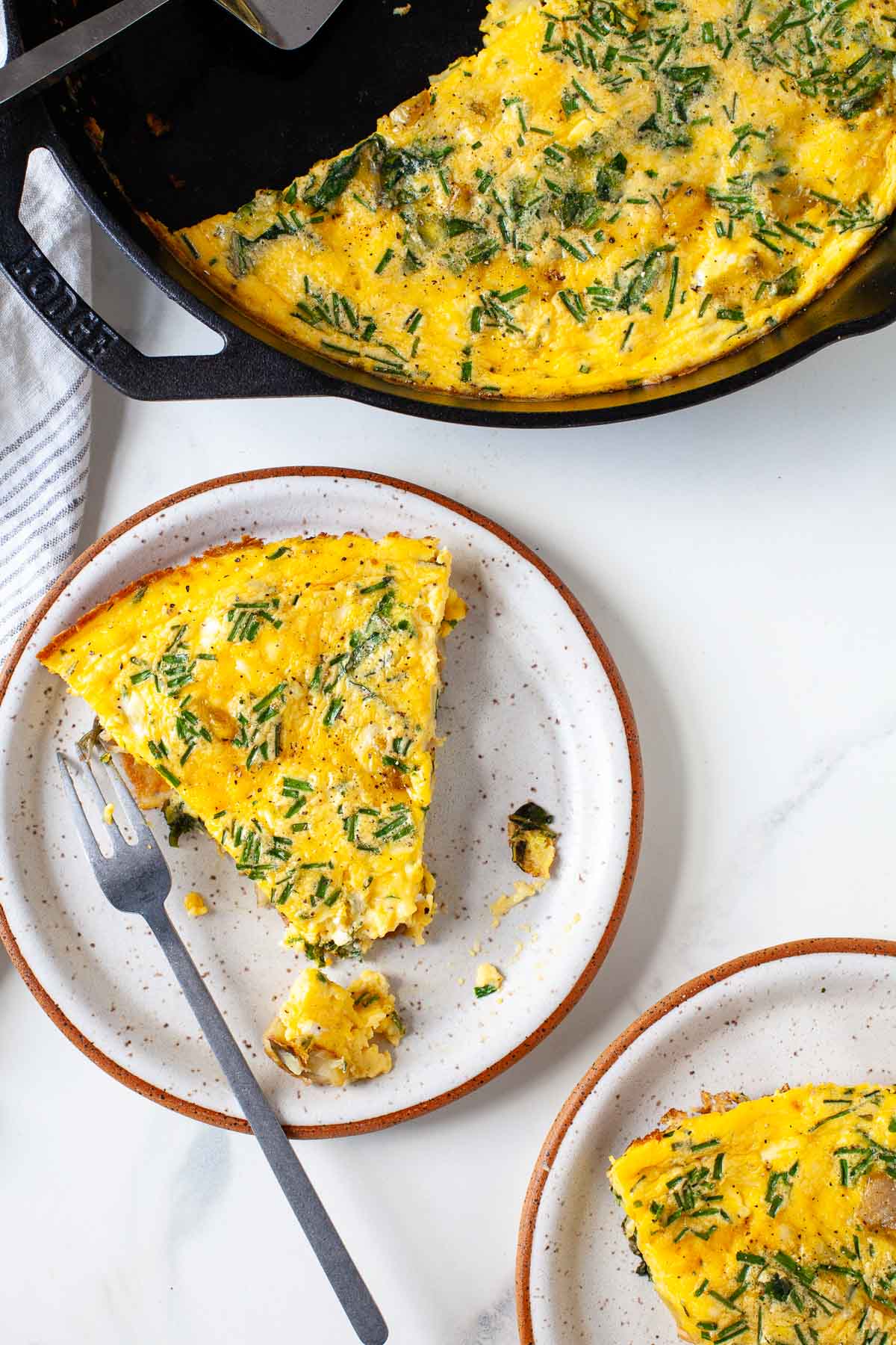 two slices of chive potato frittata on plates with fork and rest of frittata in black cast iron pan with spatula