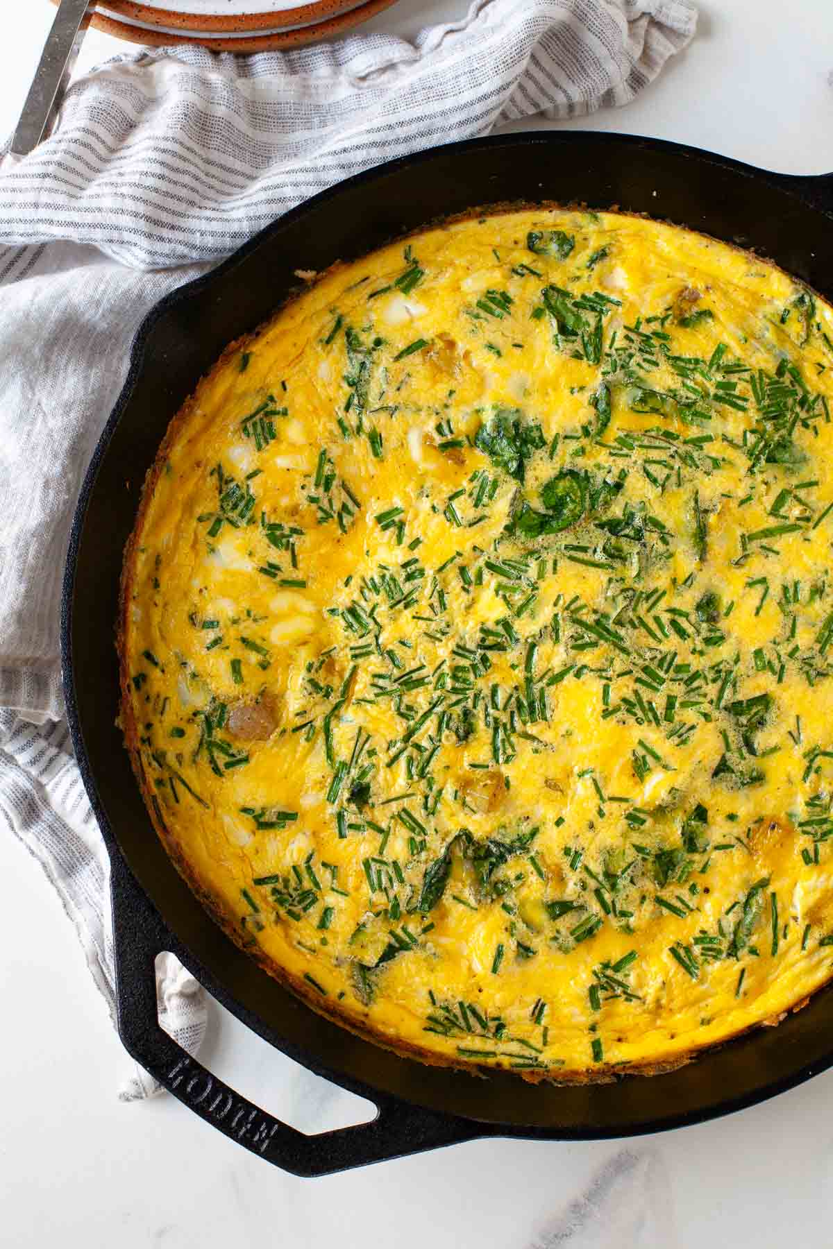 cooked frittata with chives, potato and swiss chard in black cast iron pan next to striped dish towel