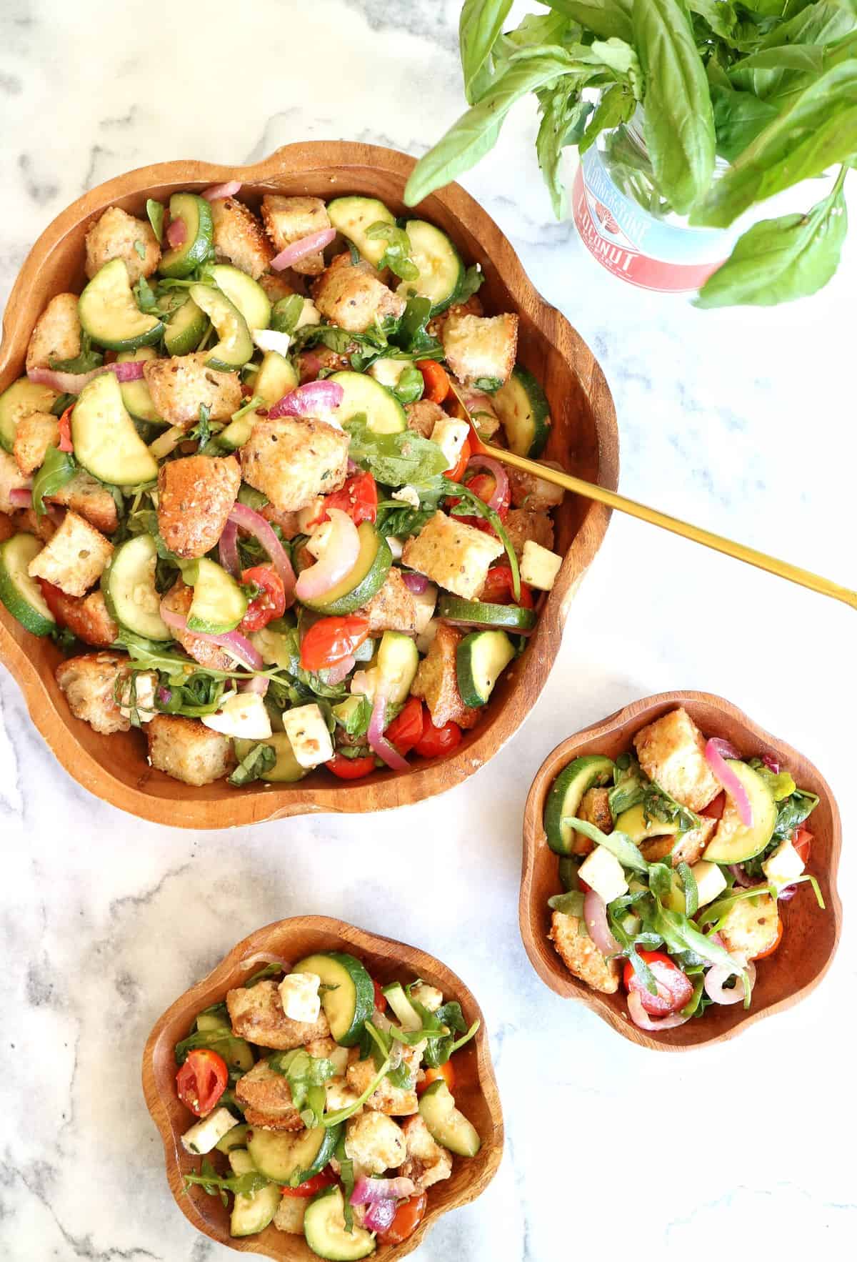 Large wooden bowl and two smaller bowls filled with roasted summer vegetable panzanella, on white marble table