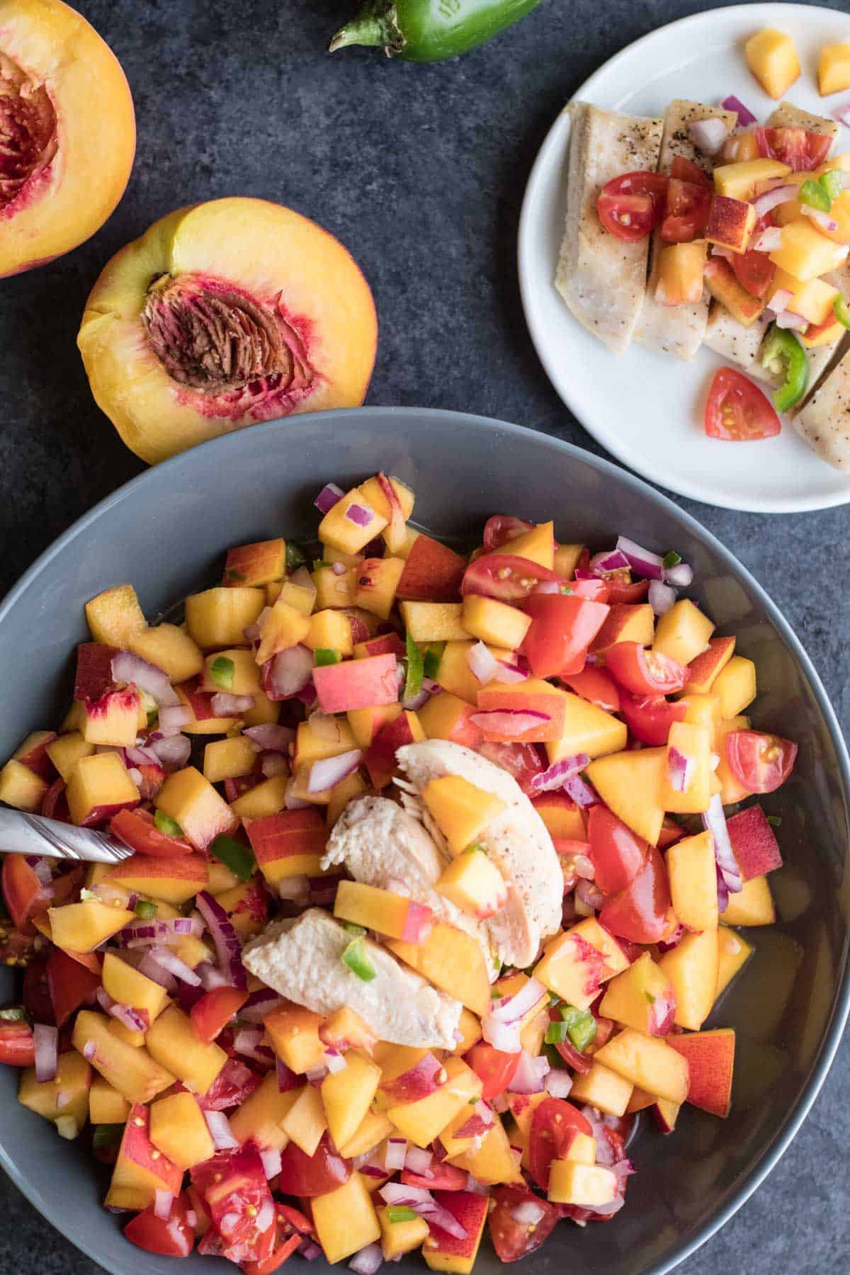 Large gray bowl with peach salsa topped with sliced chicken breast on gray slate background, peach sliced in half, small plated portion on white plate 