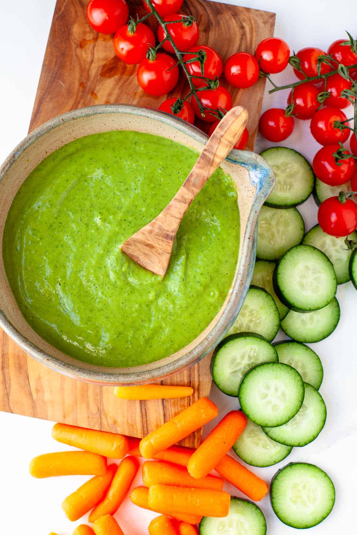 Vegan Green Goddess Dressing in a serving bowl on a wooden cutting board along with fresh cherry tomatoes, sliced ​​cucumber and baby carrots.