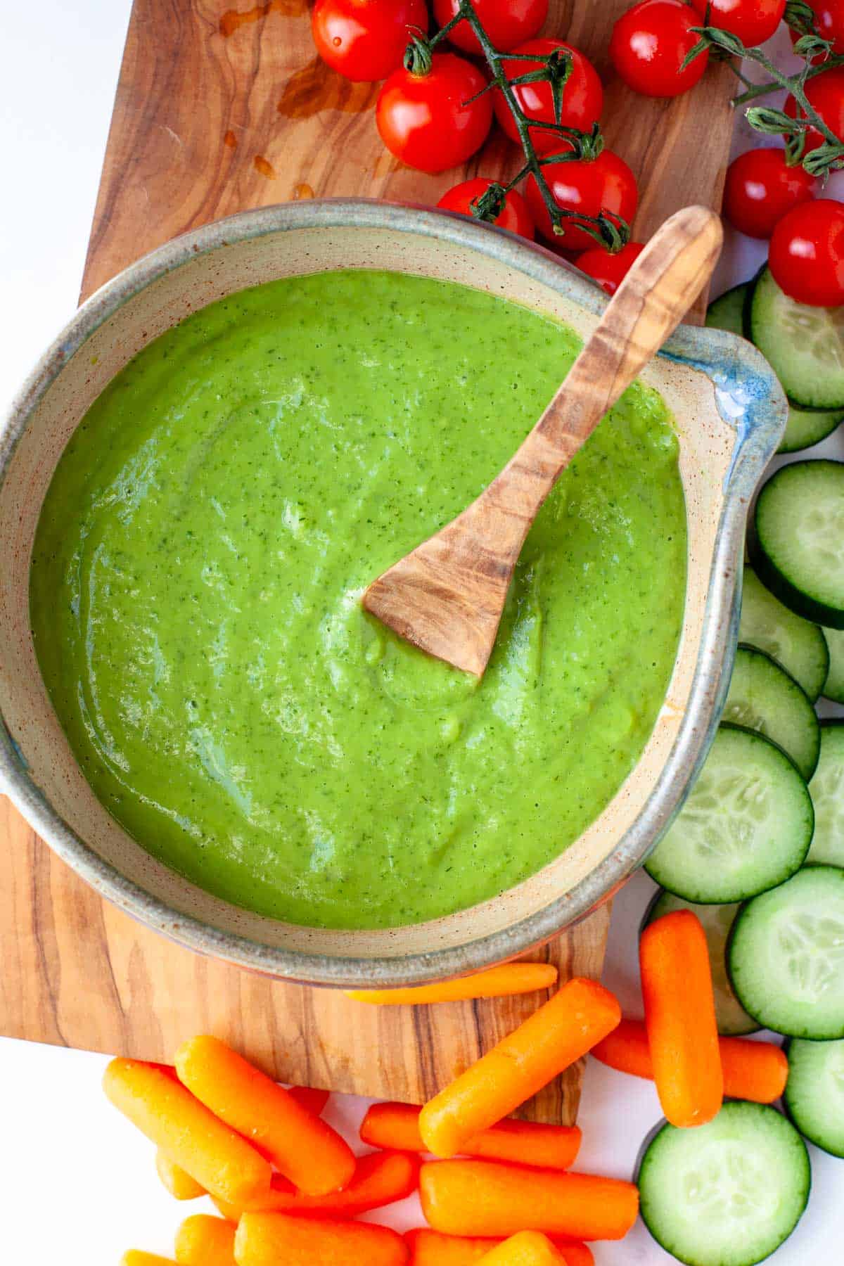 Vegan Green Goddess Dressing in a serving bowl with a wooden spoon on top of a wooden cutting board with fresh vegetables for dipping next to it.