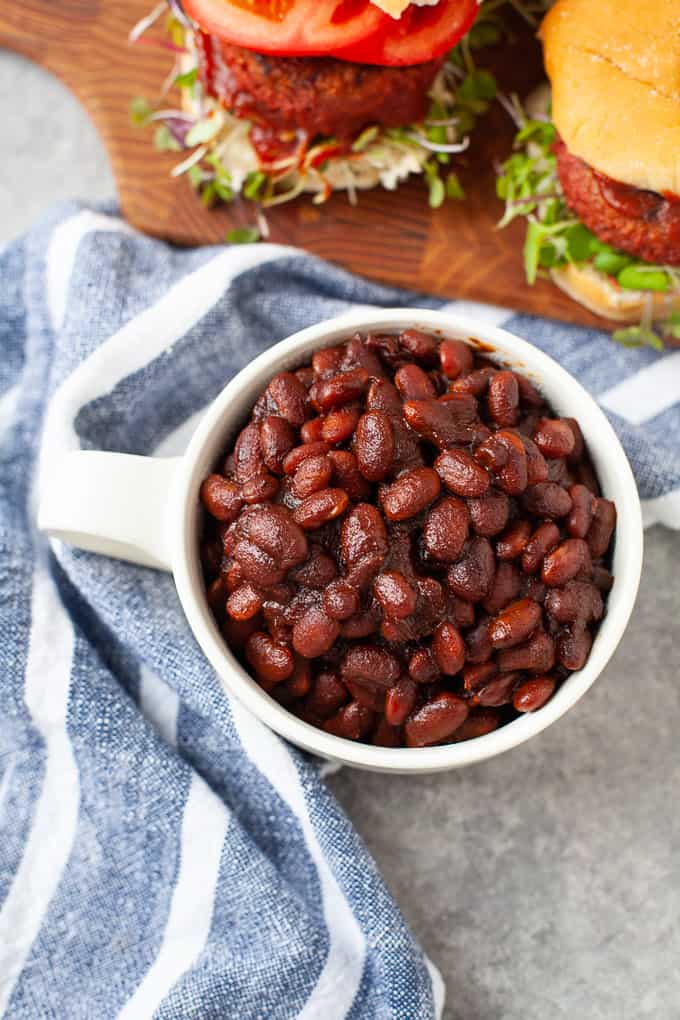 baked beans. in white mug. striped napkin. piece of burger in background.