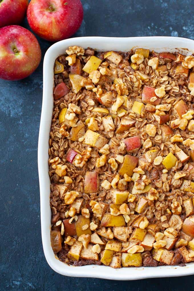 Baked oatmeal with apple pie in a white baking dish with three apples on the side