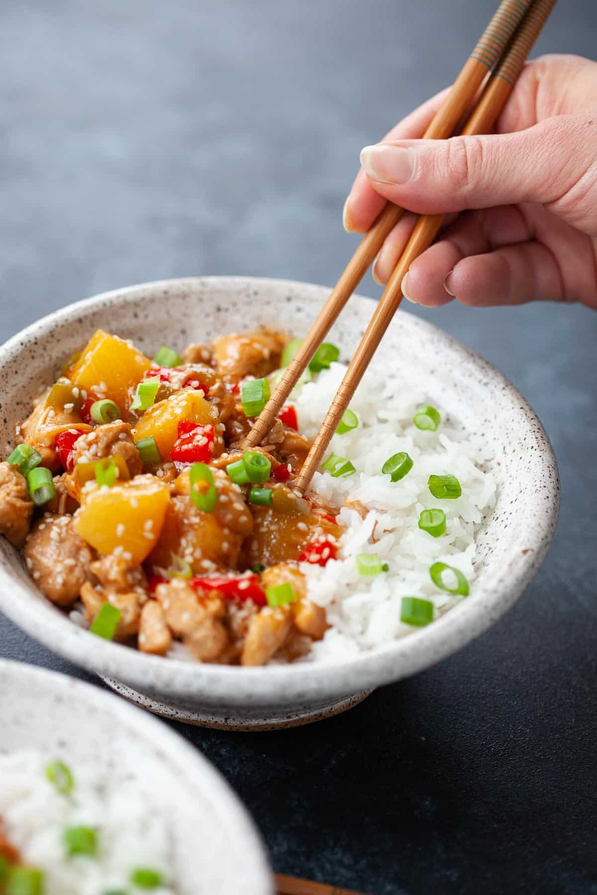 bowl of sweet and sour chicken served with white rice, green onion. hand holding chopsticks 