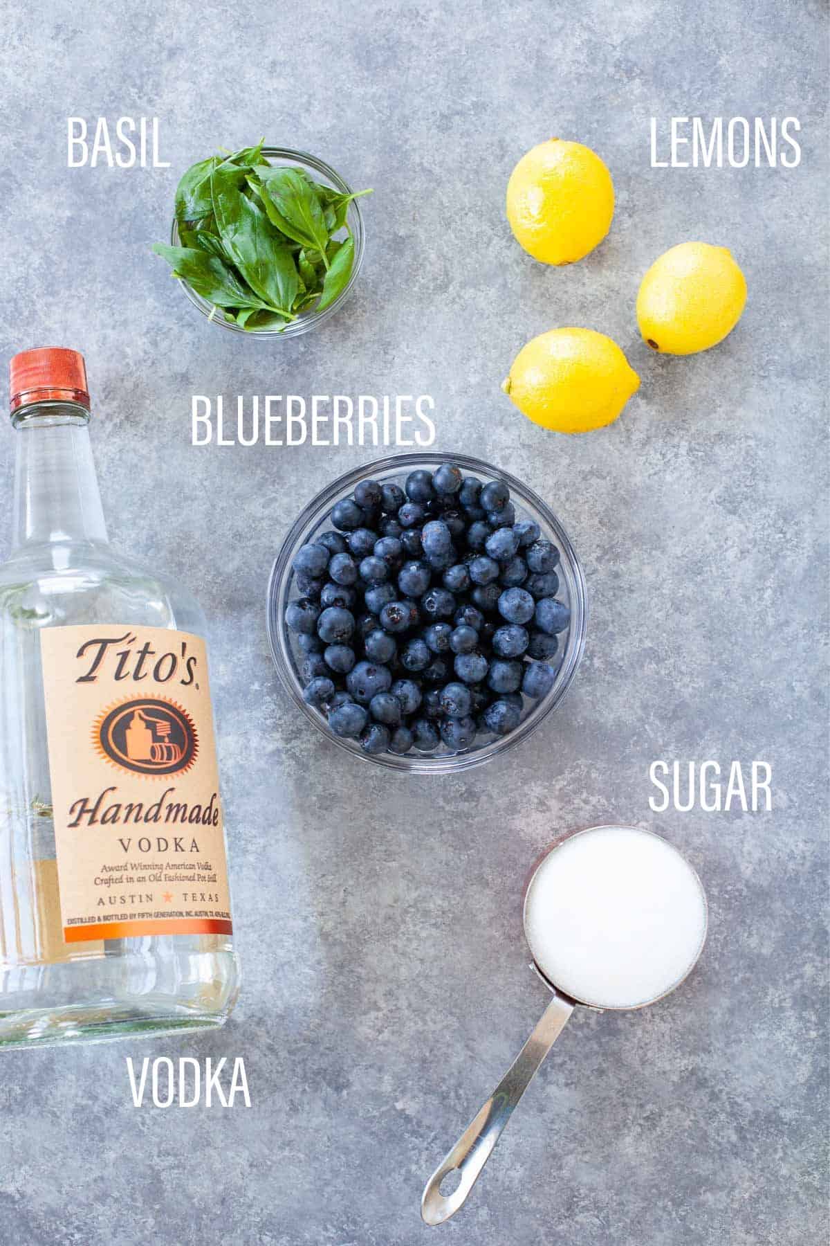 basil, lemons, bowl of blueberries, measuring cup of sugar, and bottle of vodka laid out on surface