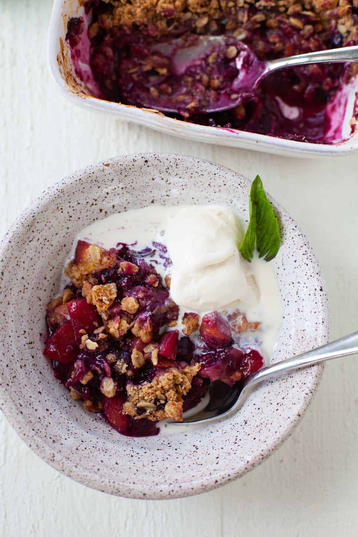 serving of Blueberry Peach Basil Crisp in a bowl with white baking dish next to it