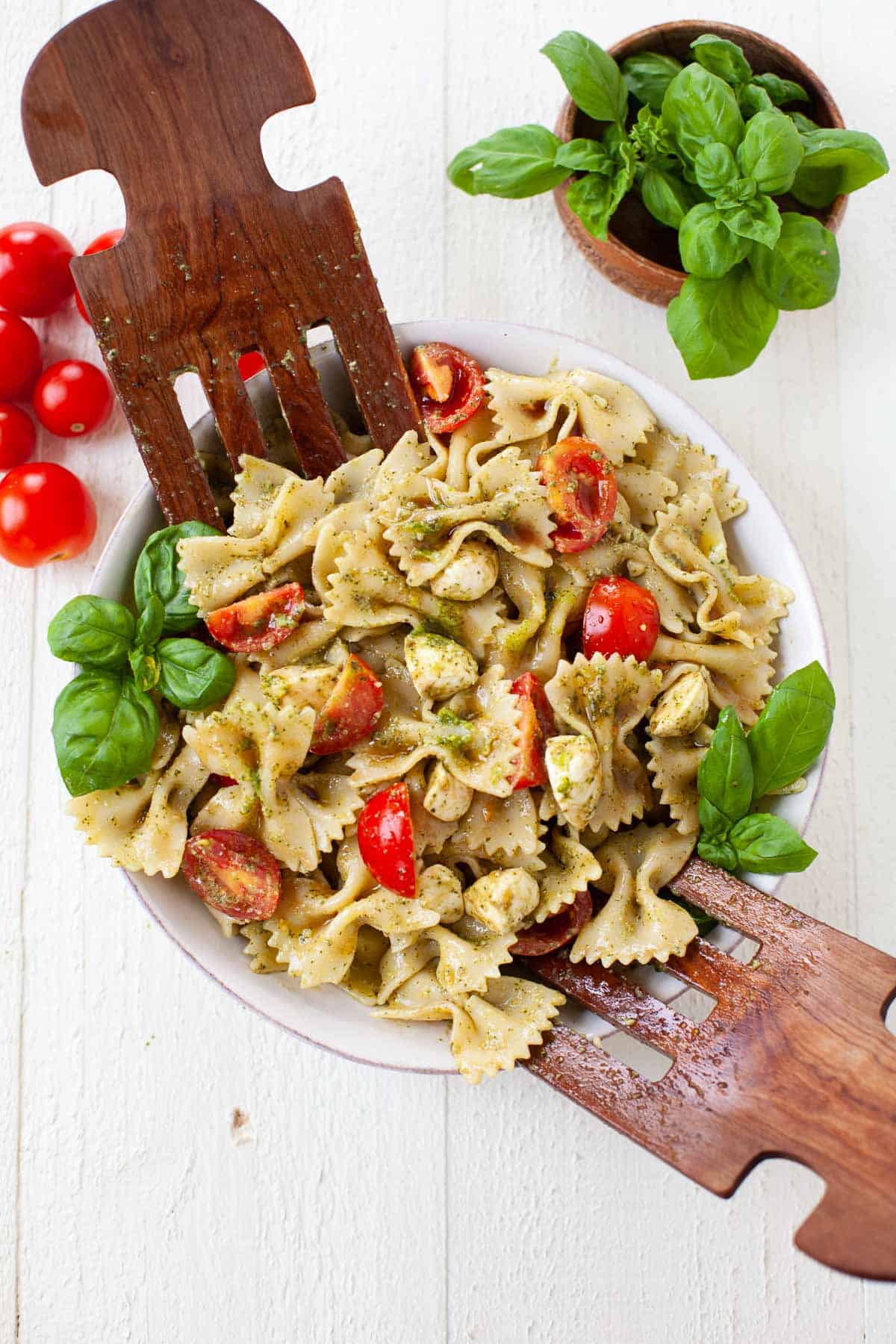 two wood spoons in a bowl of caprese pasta salad with a second bowl of fresh basil