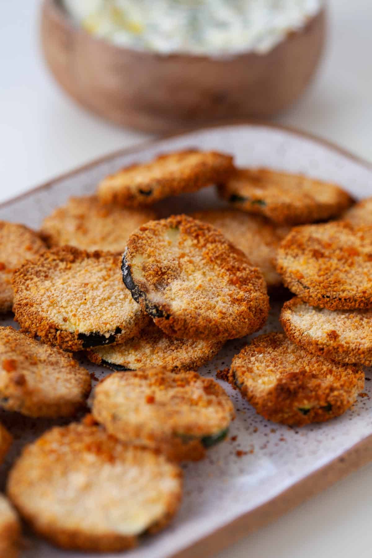 crispy zucchini chips on serving dish with dipping sauce in background