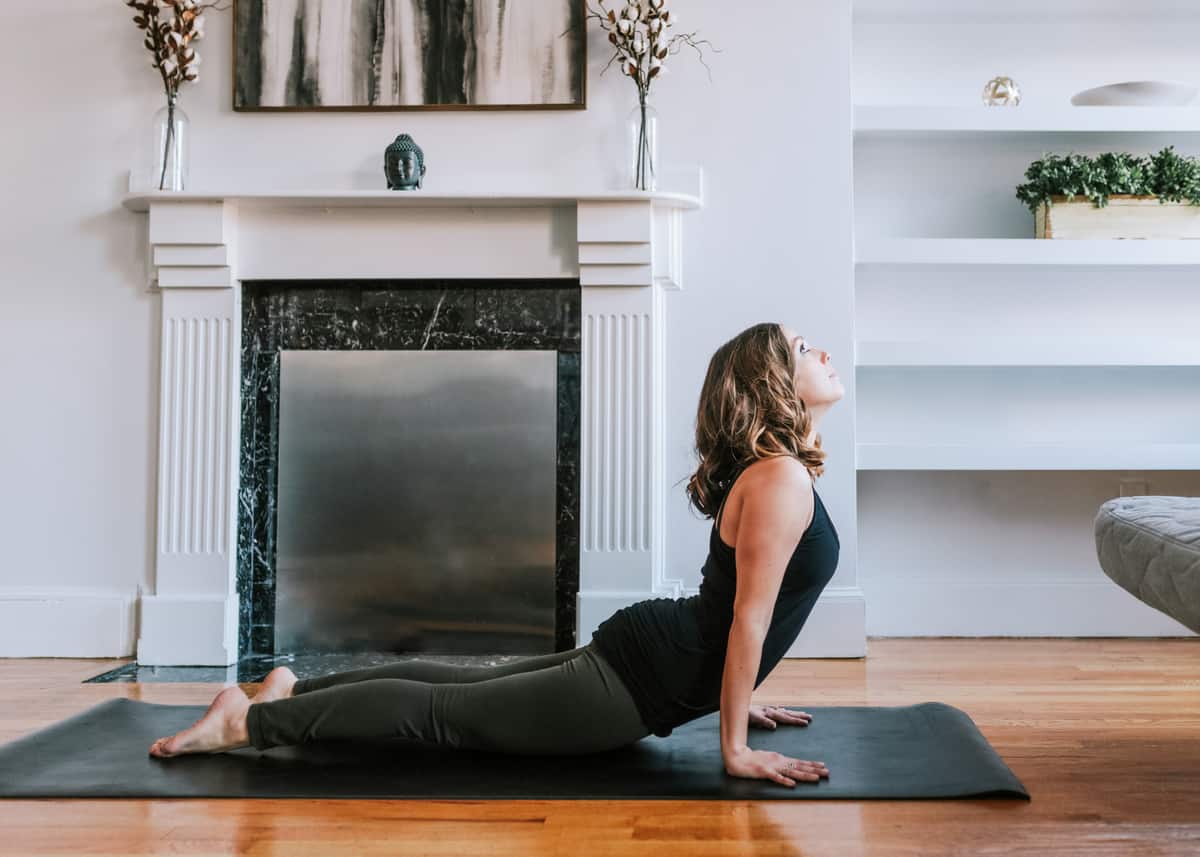 Kara is on a yoga mat demonstrating a yoga pose in front of a fireplace in her living room