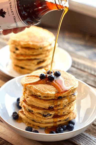 syrup being poured over a stack of fluffy Greek yogurt pancakes with fresh blueberries