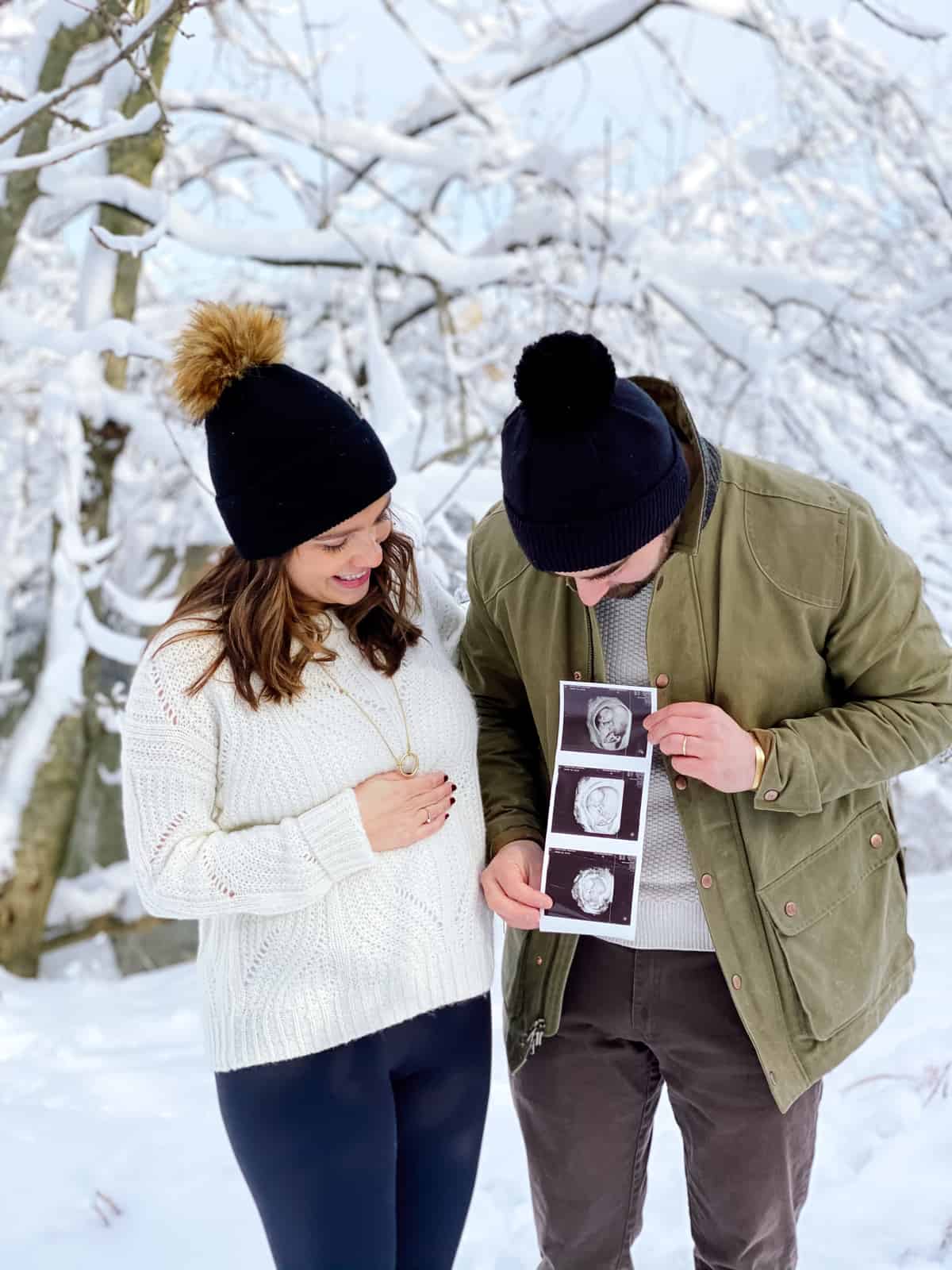 couple announcing pregnancy holding sonogram photos with snow in the background