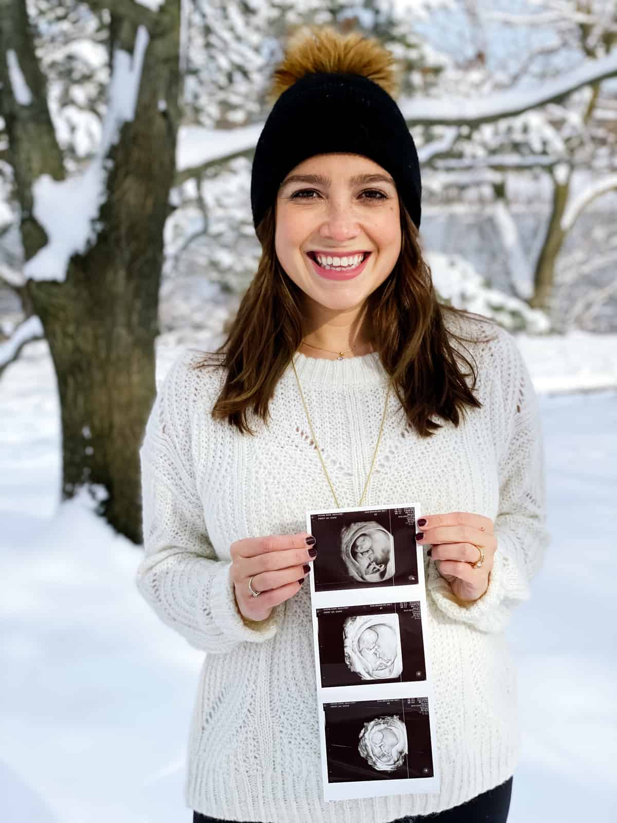 pregnant woman wearing winter hat holding ultrasound photos with snow in background