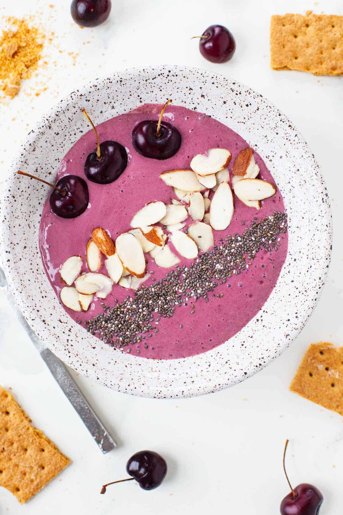 cherry cheesecake smoothie bowl with cherries, almonds, chia seeds in grey speckled bowl with graham crackers