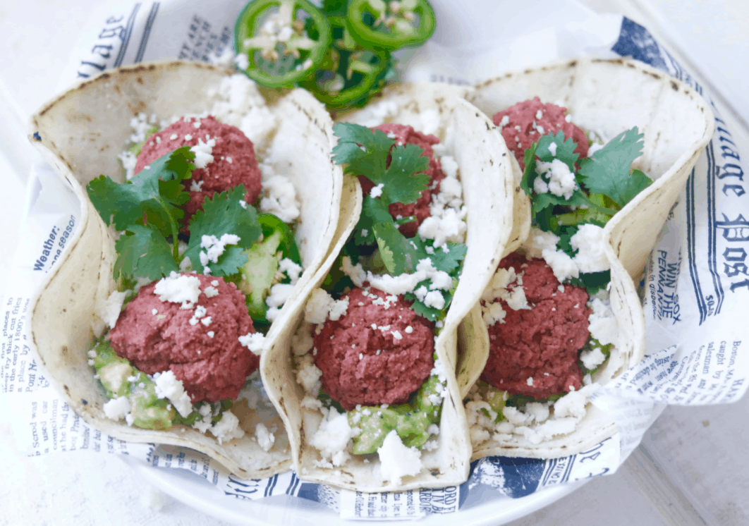 Three soft flour tacos open-face with beet falafel, crumbled vegan cheese, cucumber, and cilantro inside on a plate