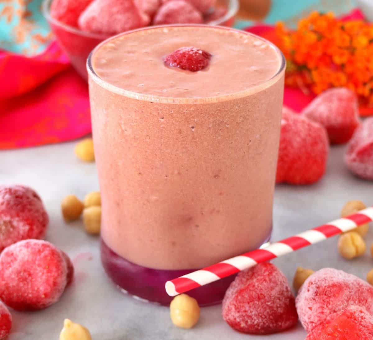 Vibrant pink smoothie in clear, short glass, strawberries and chickpeas scattered in background