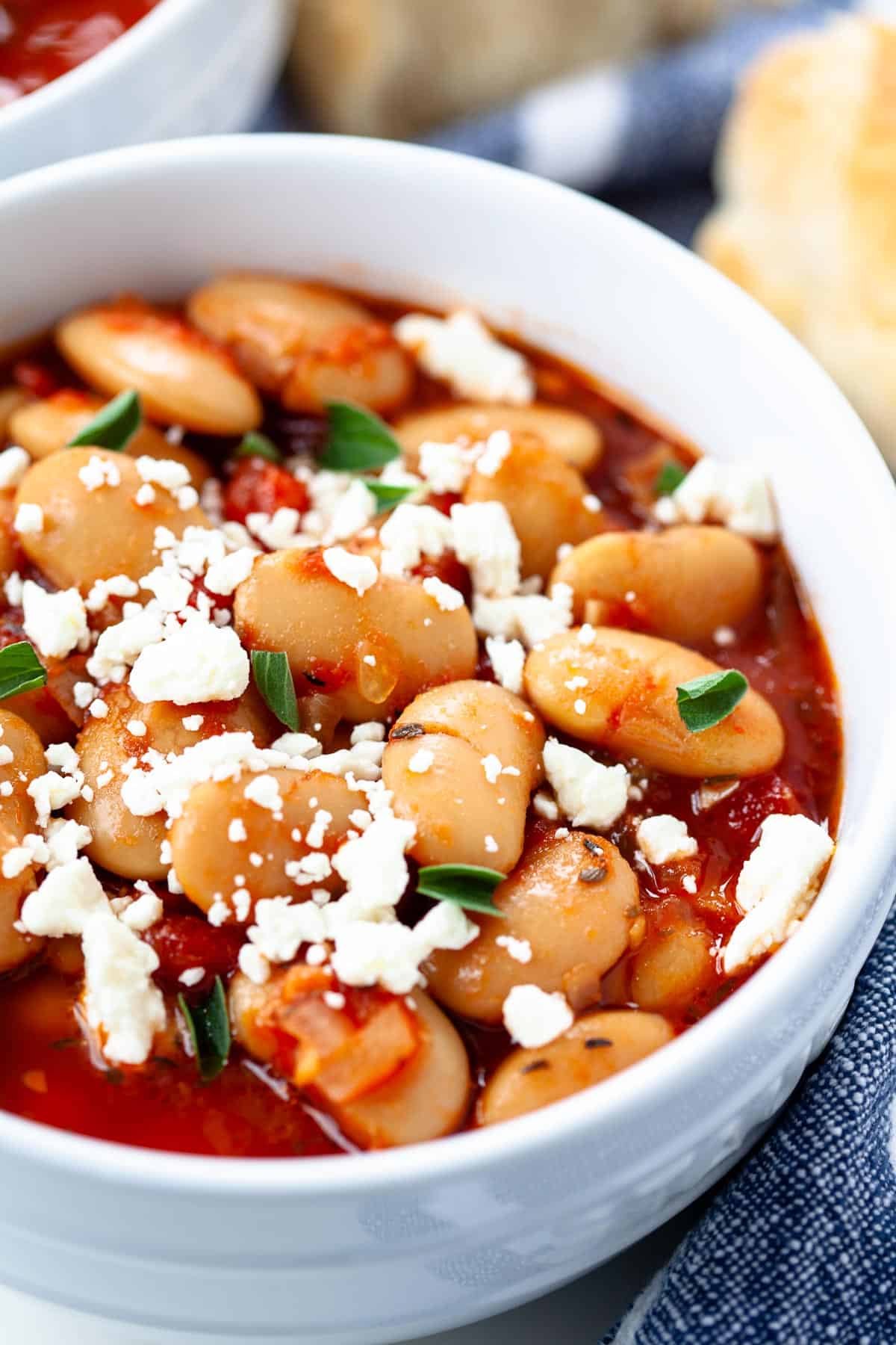 close up slow cooker greek baked beans in two white bowls feta cheese garnish blue napkin