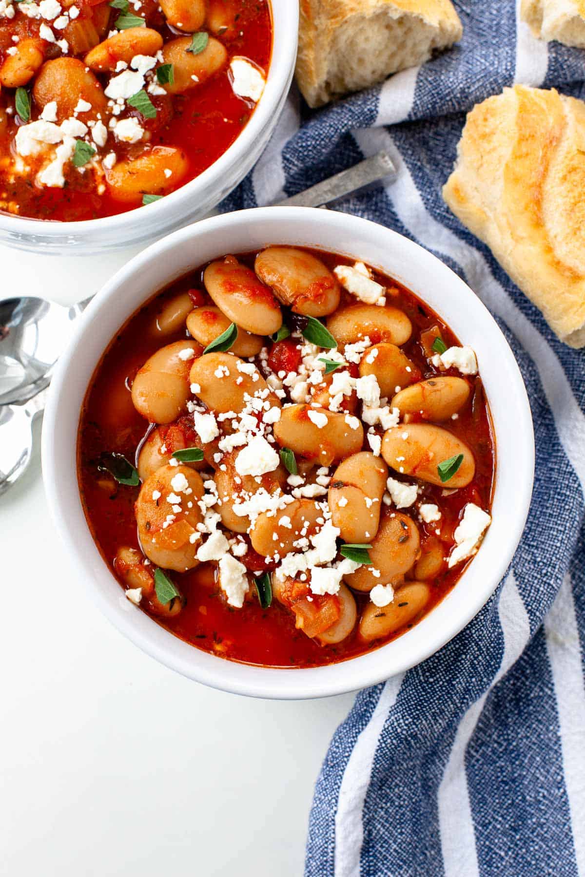 slow cooker greek baked beans in two white bowls blue napkin bread and spoons background