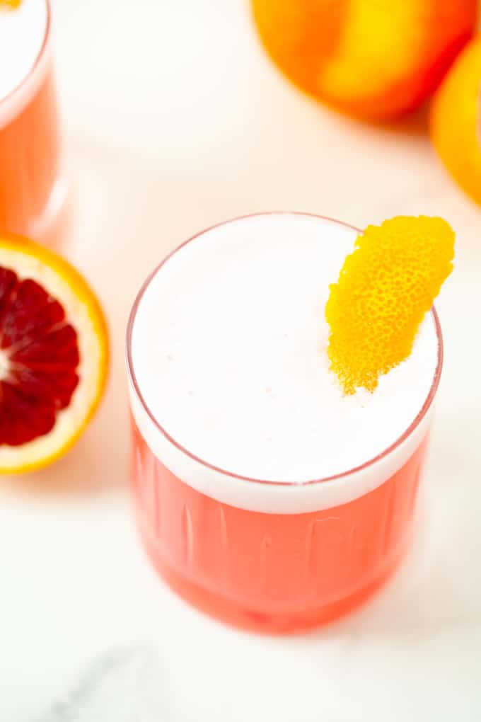 Overhead image of pink cocktail on white marble, white foam, orange rind garnish