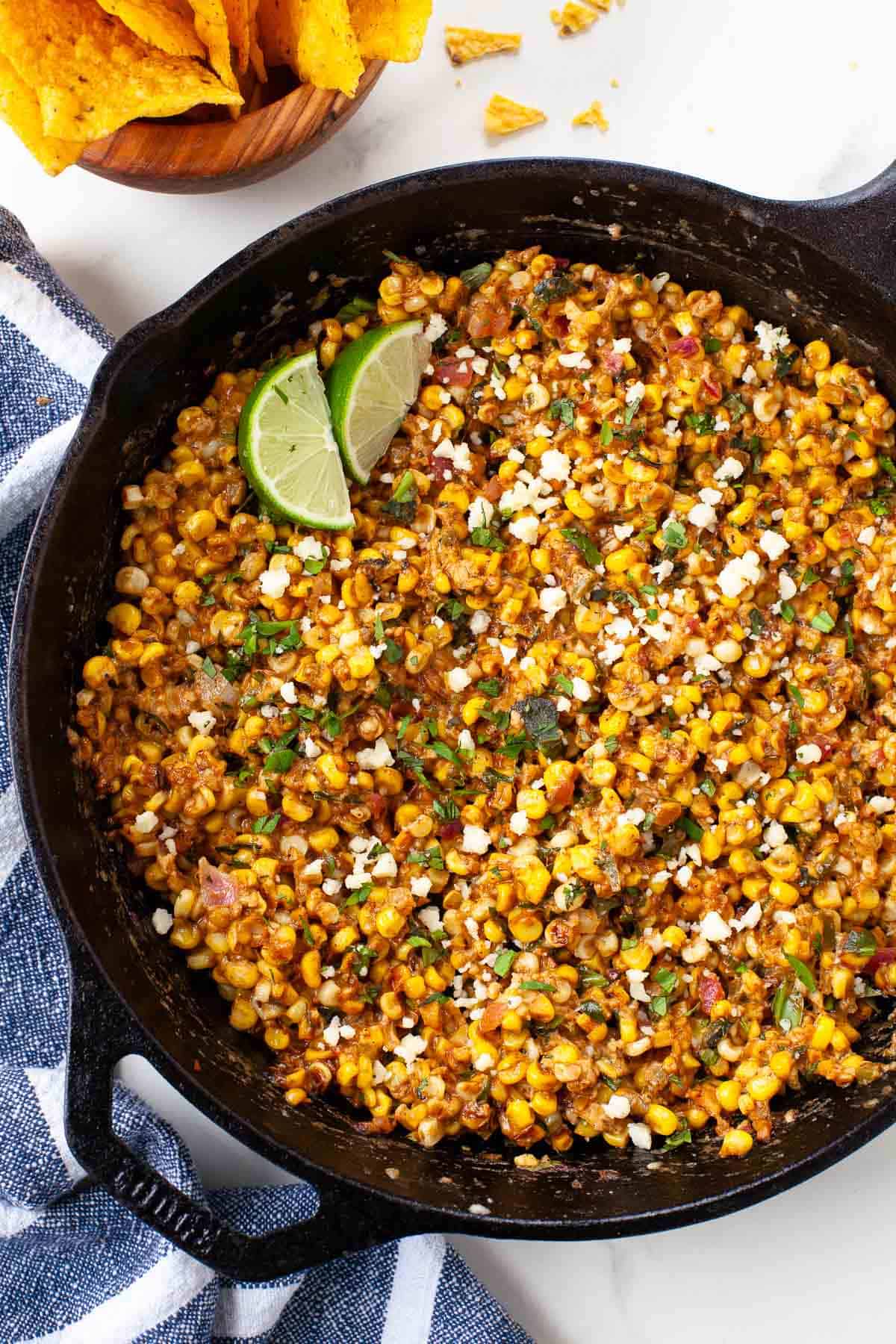 mexican street corn dip in cast iron with limes, cilantro and small wood bowl with chips