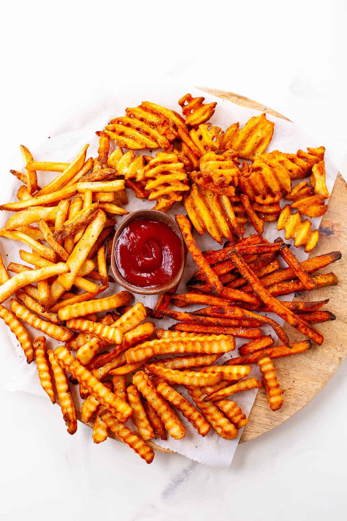 crispy crinkle, waffle, sweet potato, regular fries on circular wooden plate. white napkin underneath. small wooden bowl with ketchup. on white marble counter.