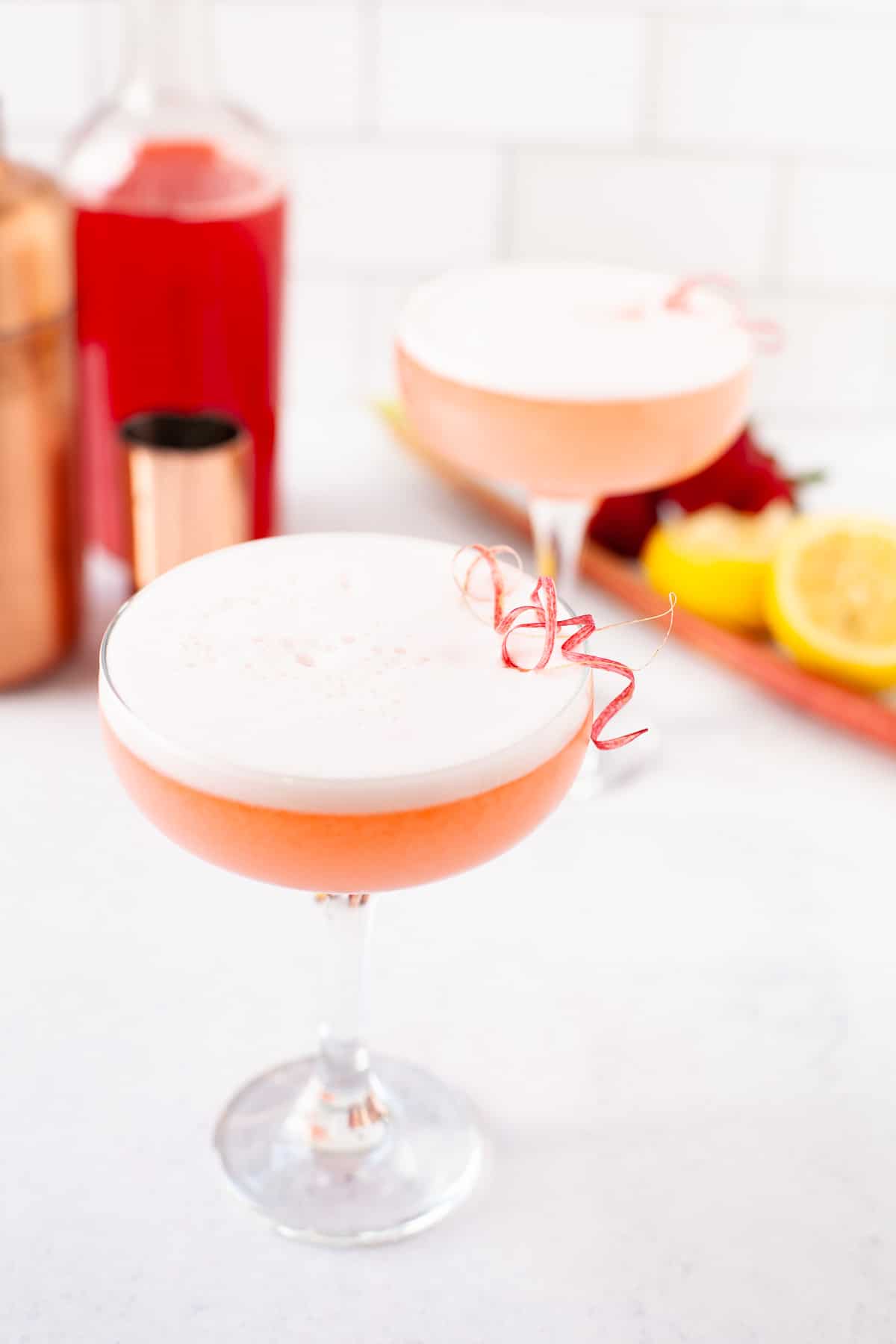 two strawberry rhubarb cocktails in coupe glasses with rhubarb peel garnishes, lemons and syrup in background