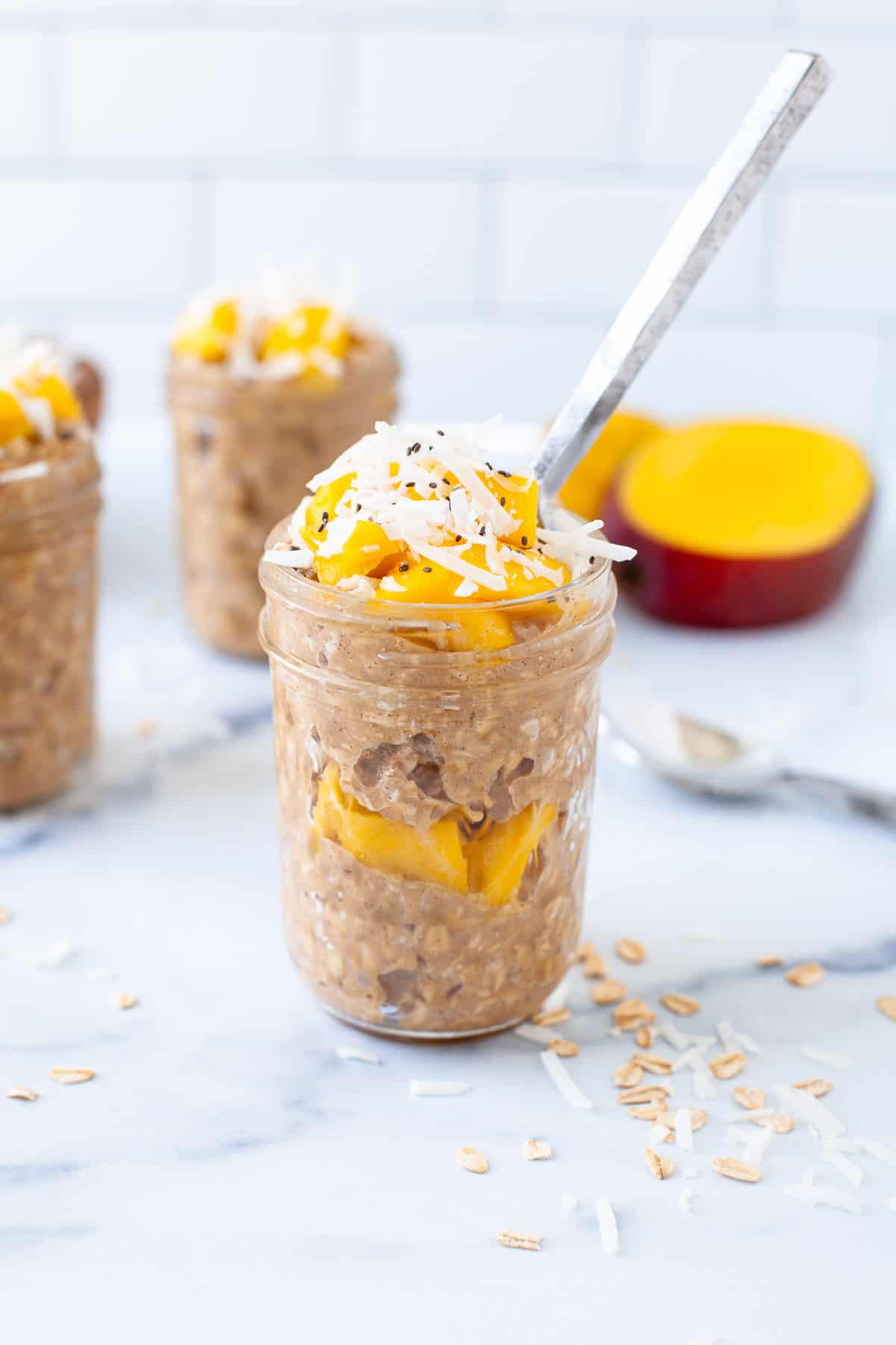 3 mason jars overnight oats. two in background. one with spoon. layer of mangos. topped with mangos. coconut shreds. chia seeds. spoon. sliced mango. 
