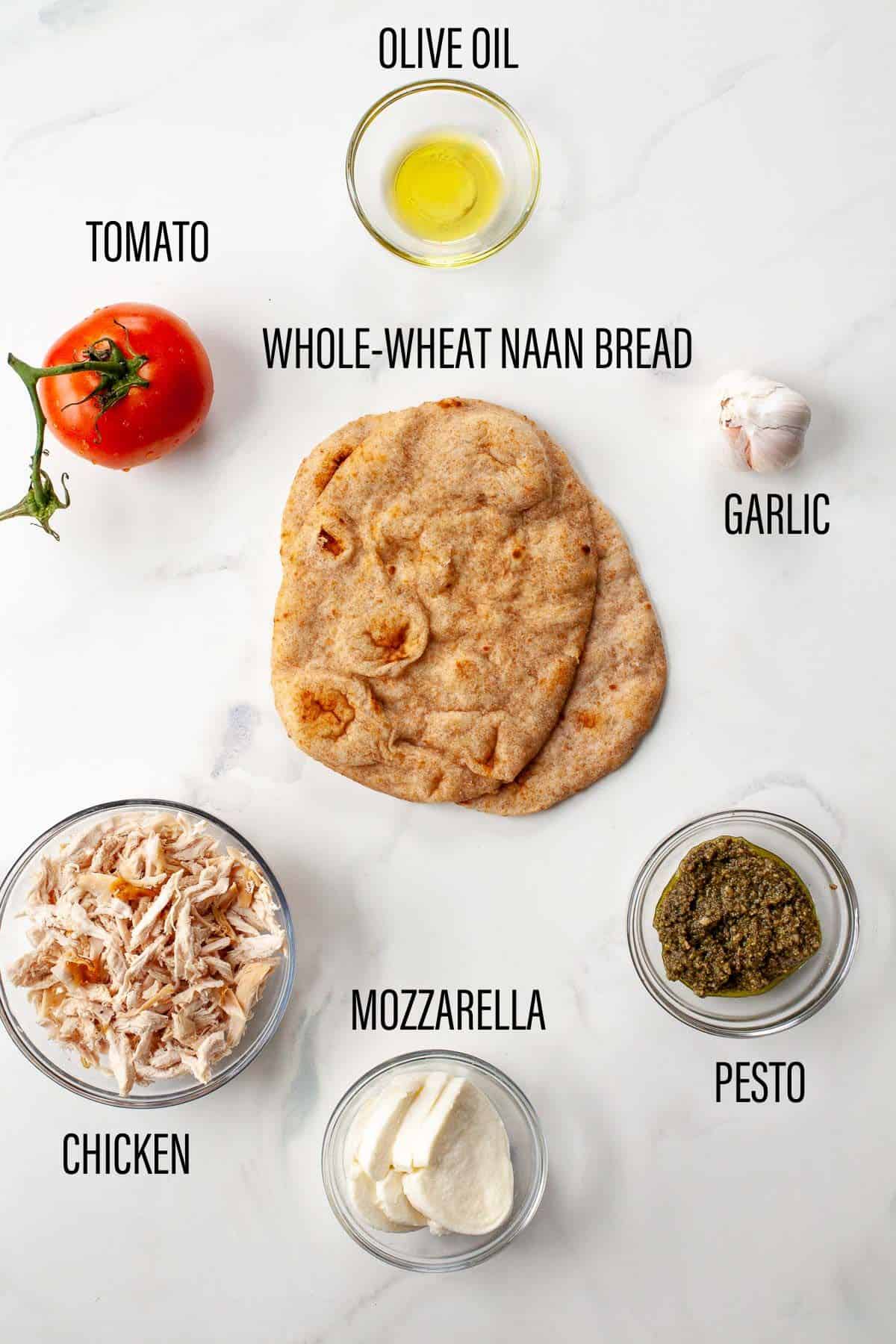 ingredients for flatbread on marble surface tomato, olive oil, naan bread, garlic, chicken, mozzarella, pesto