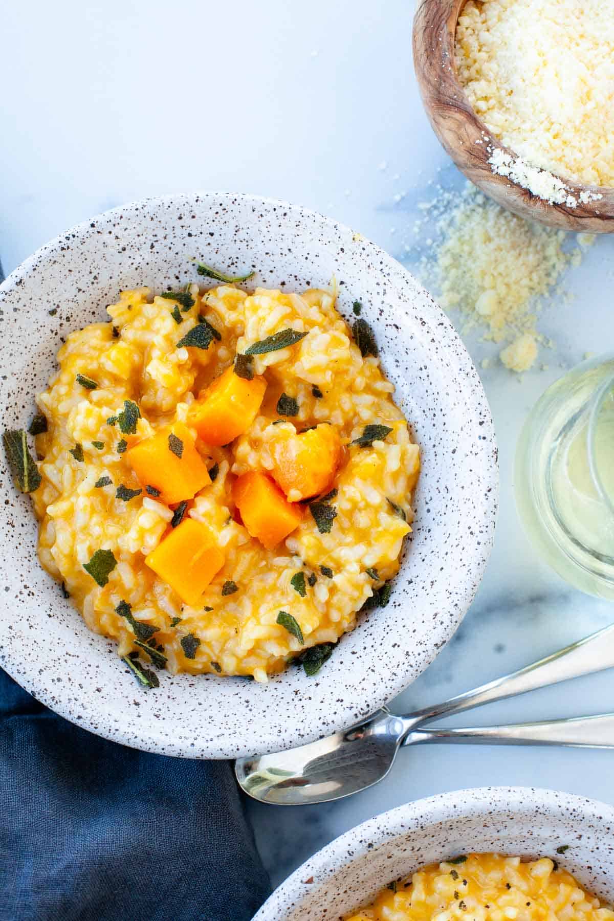 butternut squash risotto in grey speckled bowl, two spoons, glass of wine, and parmesan cheese