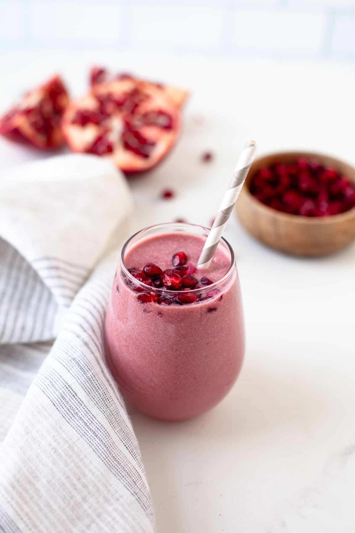 Pink smoothie in a glass with pomegranate seeds and a straw. A whole pomegranate and bowl of seeds in the background.