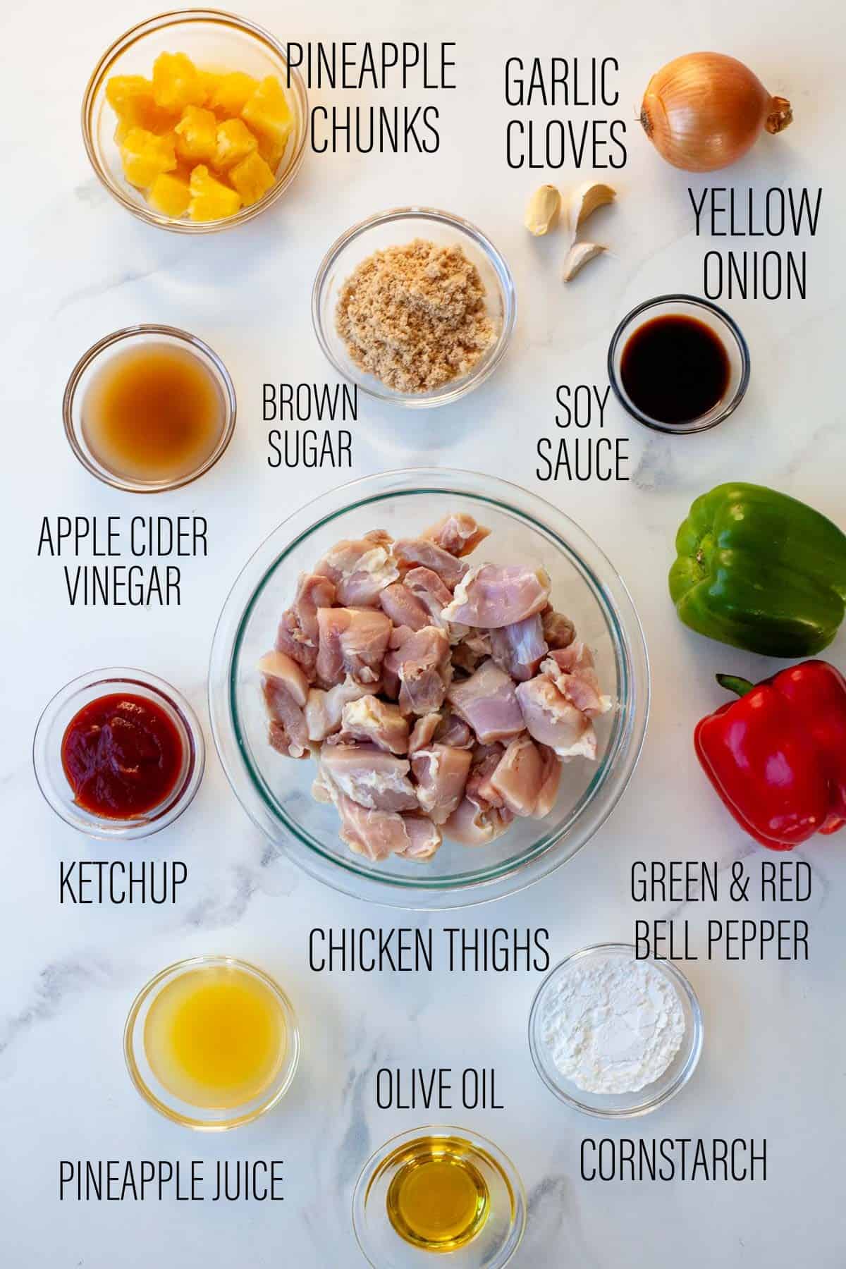 Instant pot sweet and sour chicken ingredients shown. Chicken thighs in the center surrounded by other ingredients in small bowls. Pineapple chunks, garlic cloves, yellow onion, soy sauce, brown sugar, apple cider vinegar, ketchup, green and red bell pepper, olive oil, pineapple juice, cornstarch
