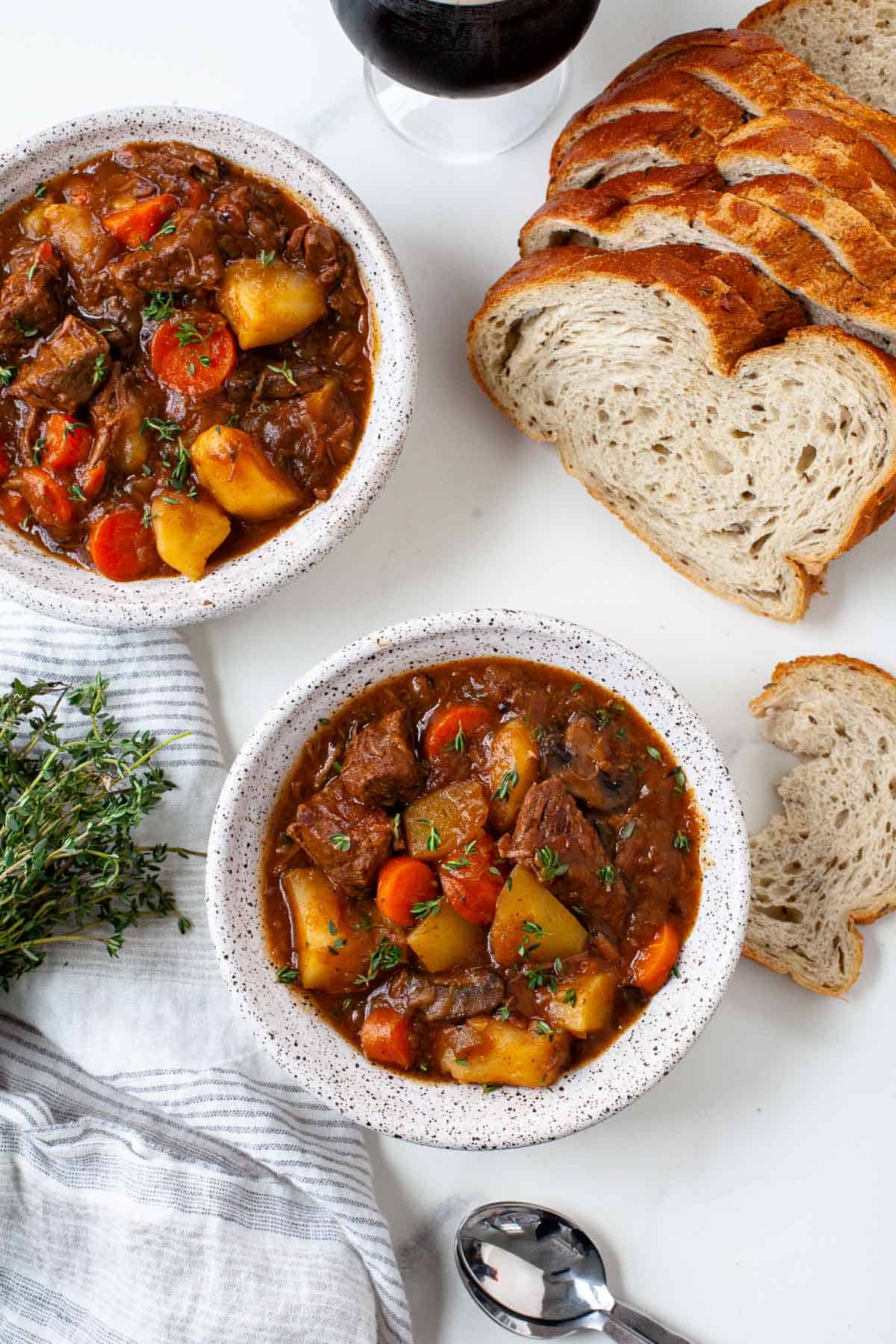 beef stew with potatoes and carrots in white speckled bowls with silver spoons, loaf of bread, striped towel