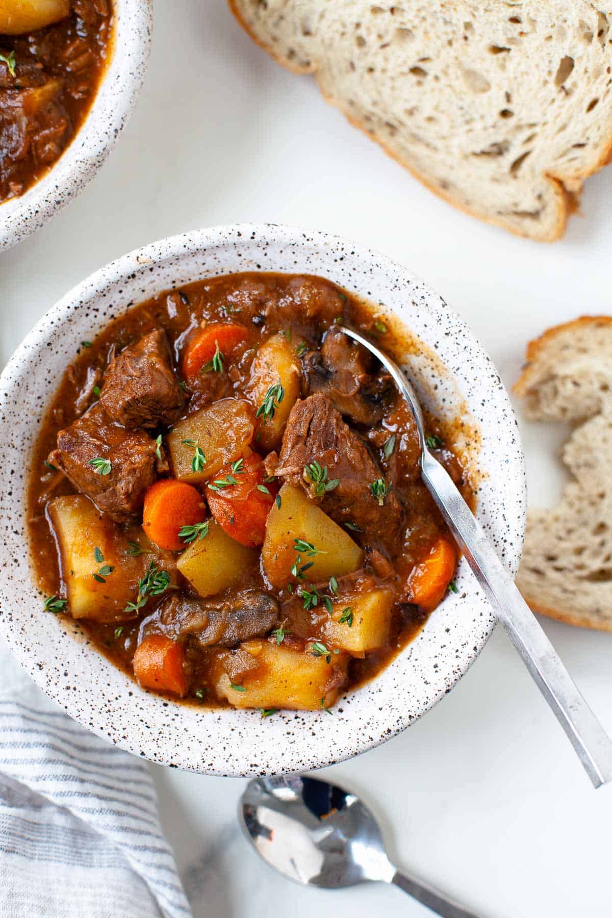 beef stew with potatoes and carrots in white speckled bowl with silver spoon, two slices bread.