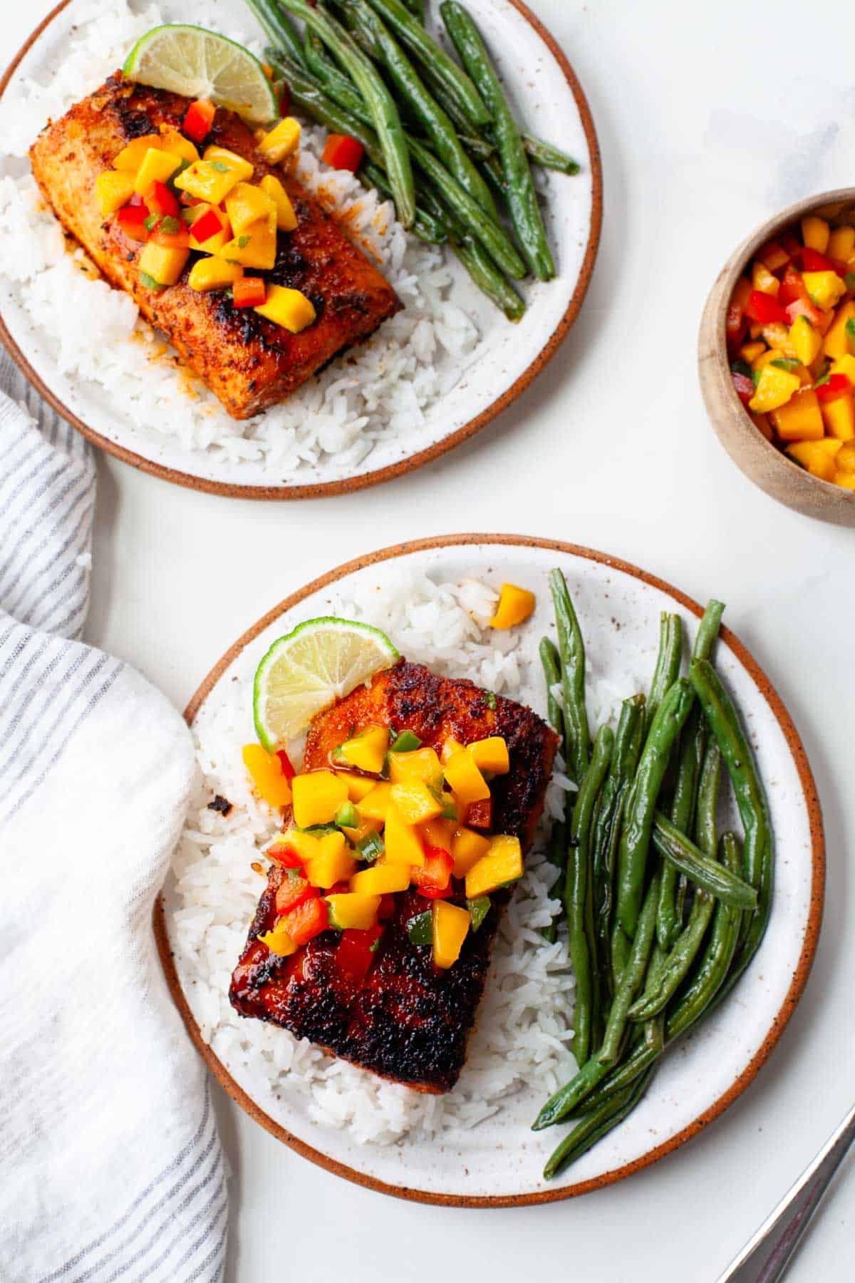 two plates of blackened mahi mahi on beds of white rice, garnished with fresh mango salsa and served with green beans