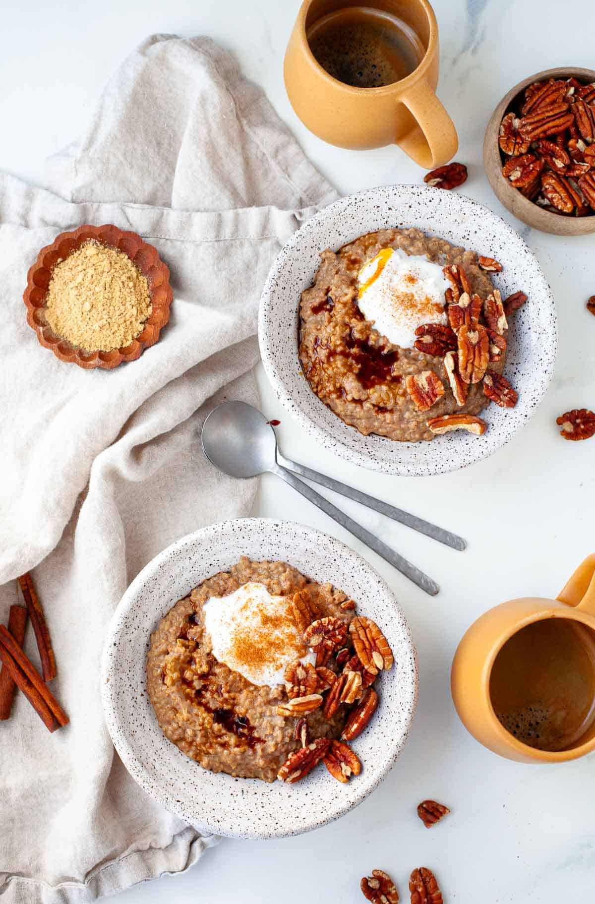 Two white speckled bowls filled with oat gingerbread with yogurt, pecans and cinnamon next to two orange coffee mugs, a small bowl of pecans and a kitchen towel