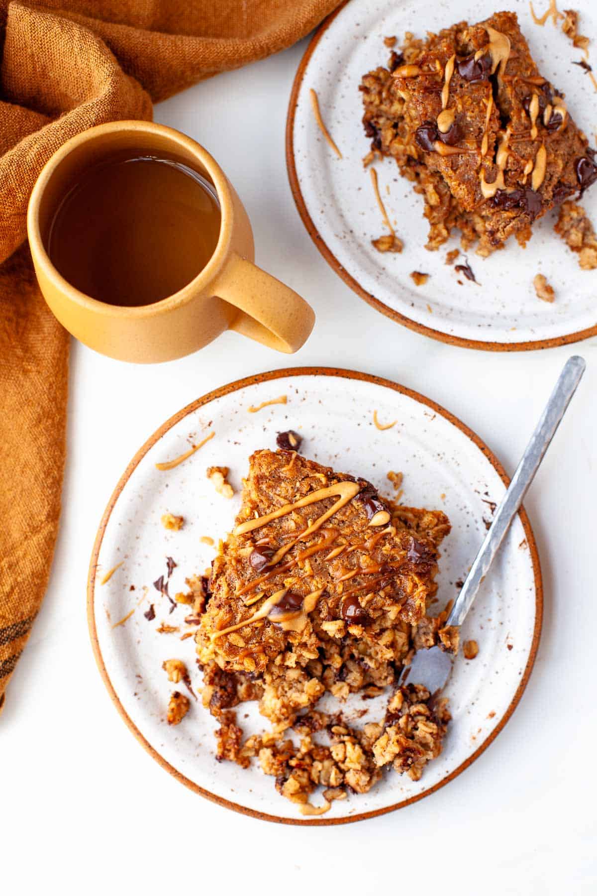 two white speckled plates with peanut butter baked oatmeal drizzled with peanut butter and topped with chocolate chips, beside orange coffee mug