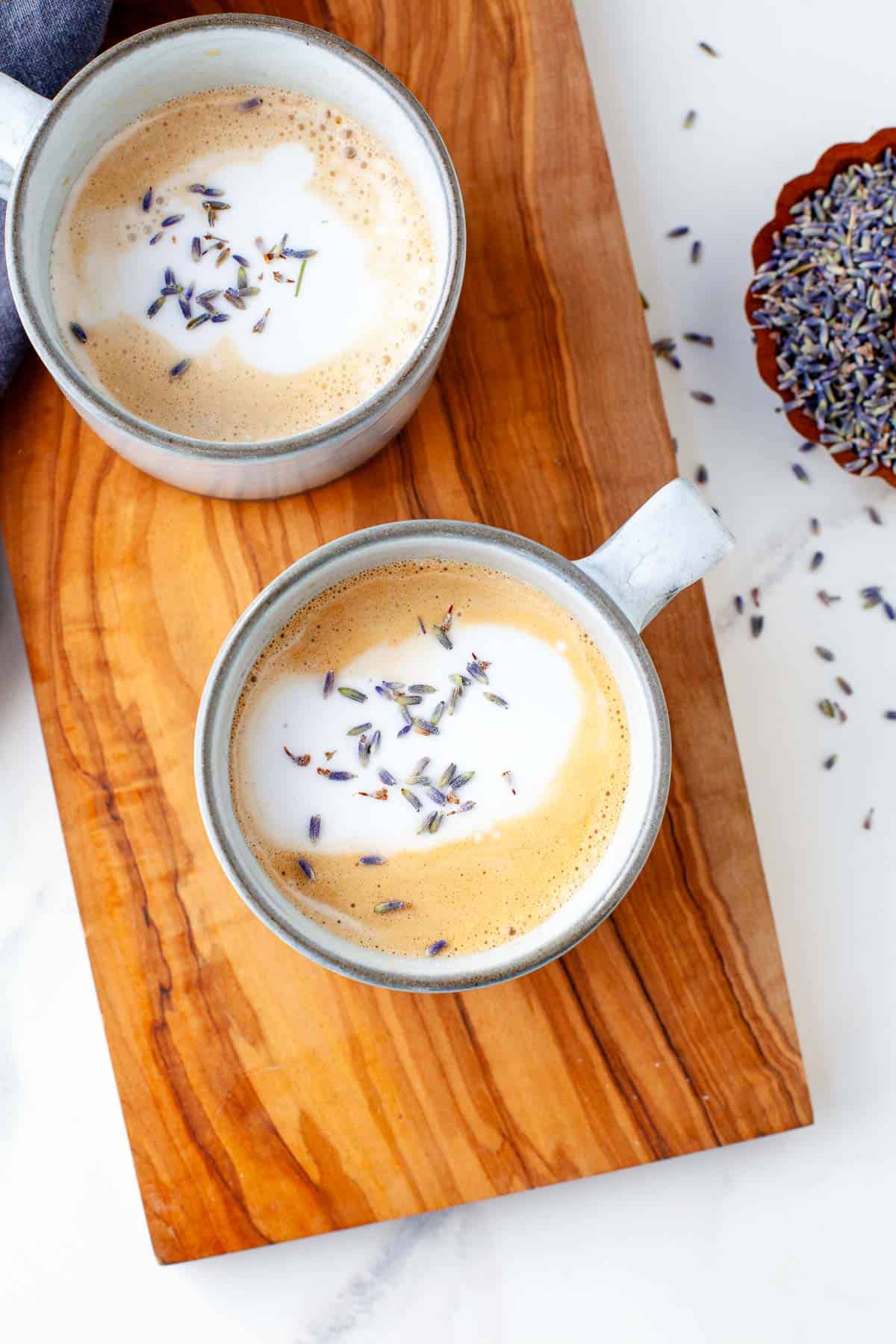two lavender lattes in white mugs garnished with lavender buds atop wooden surface.