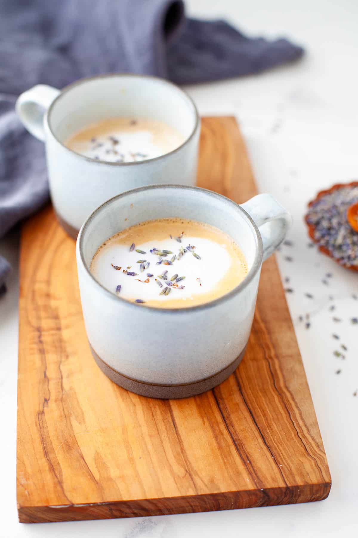 two lavender lattes in grey mugs on wood board topped with foam and garnished with lavender buds 
