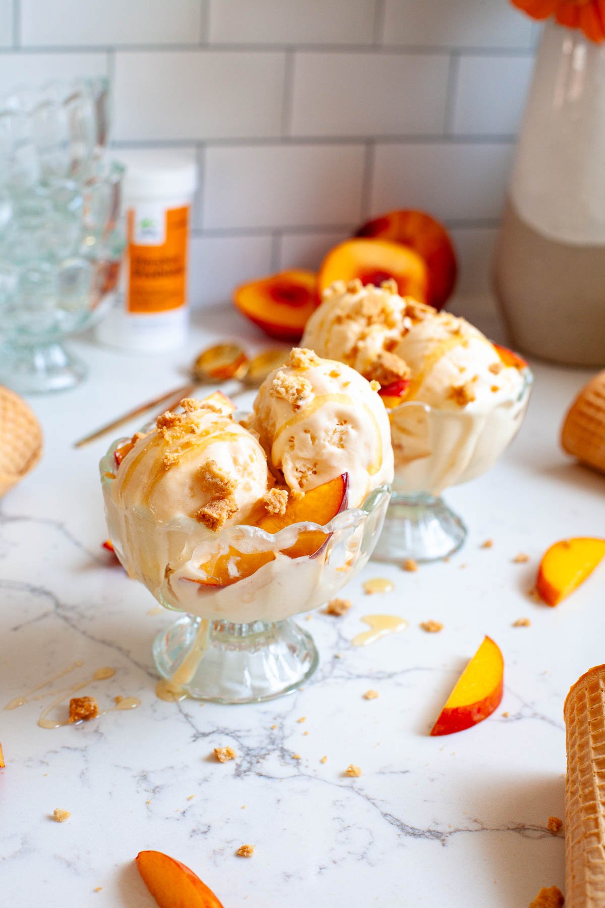 two glass sundae bowls with peach ice cream topped with honey, cookie crumbs and peach slices