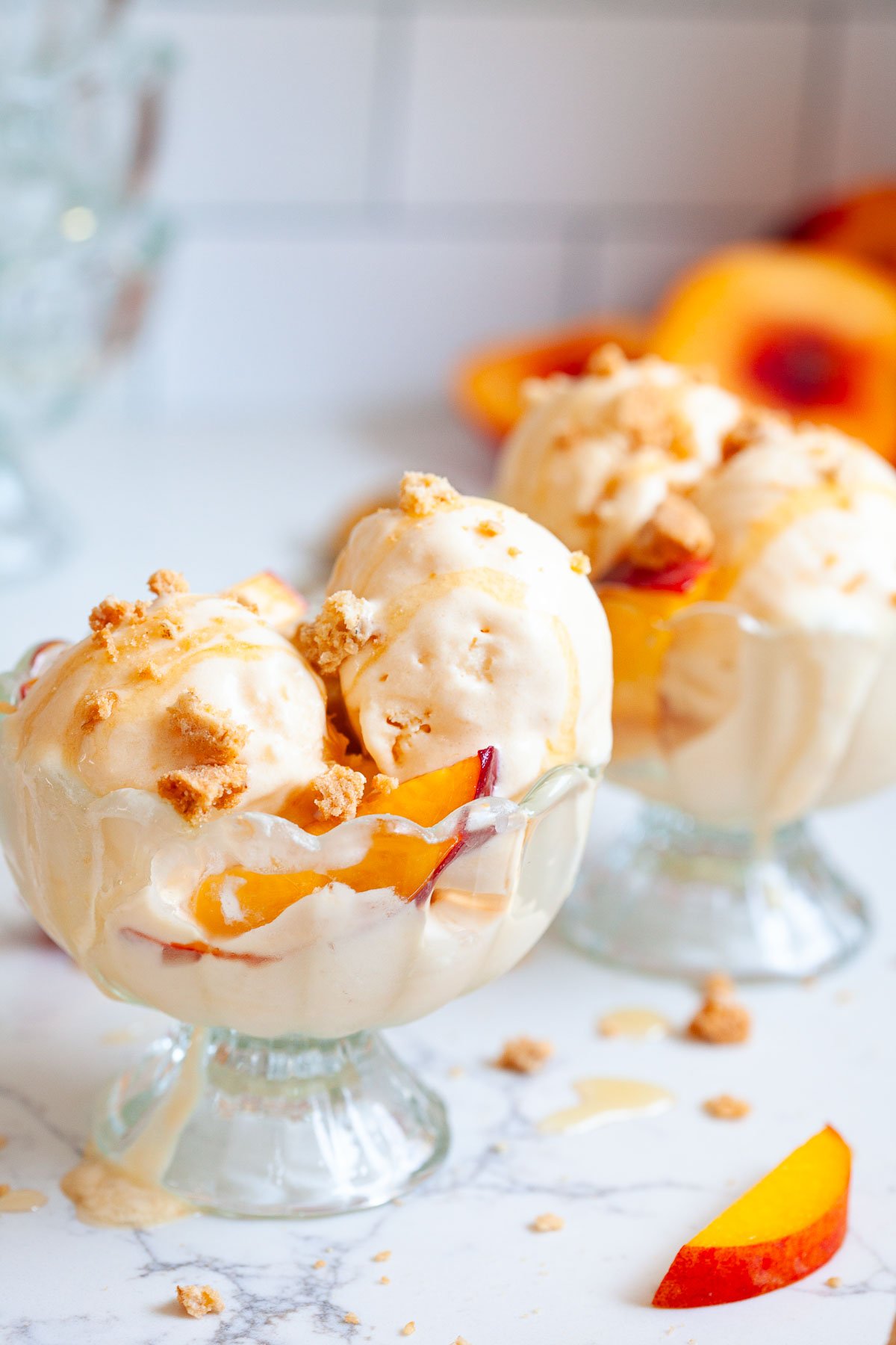 two glass sundae bowls with scoops of peach ice cream with peach slices and cookie crumbles