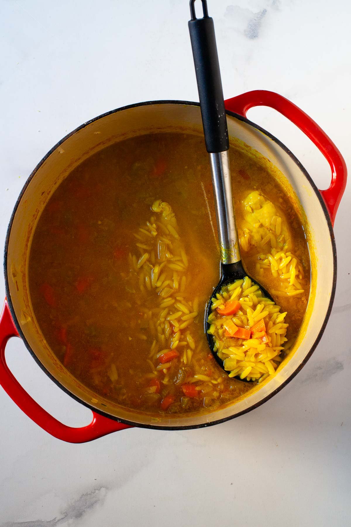 cooked chicken soup with orzo in red dutch oven with black mixing spoon on marble surface